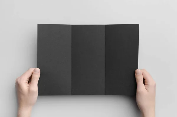 Black A4 Tri-Fold Brochure Mock-Up - Male hands holding a black tri-fold on a gray background.