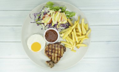 Beyaz ahşap zemin masa sahne dekorasyonu baharatlı üzerinde beyaz bir tabak içinde domuz eti biftek sosu ve mor karnabahar, Top görünümü kızarmış yumurta ile kızarmış patates, yeşil salata,.