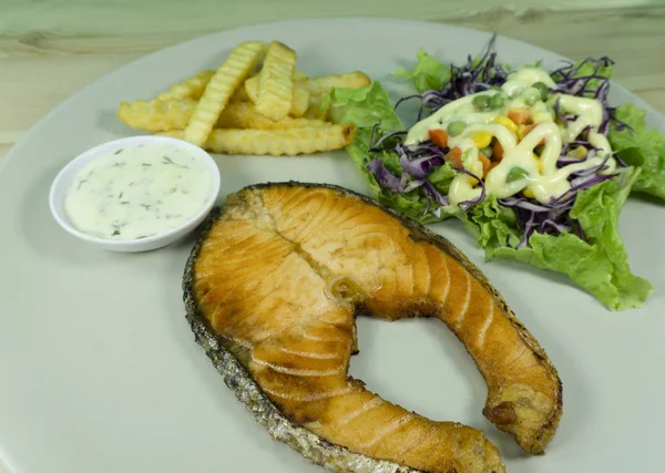 Salmon steak with tartar sauce in a white dish on a wooden background decoration French fries, green salad, purple cauliflower,Top view