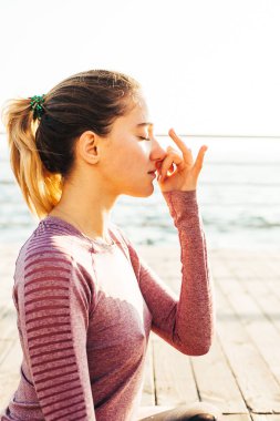 Genç bir kadın şafak vakti deniz kıyısında pranayama yapıyor.