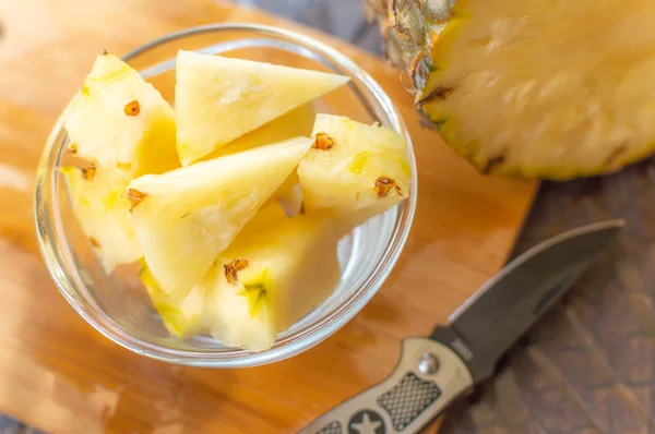 Close up of pineapple pieces and slices kept in glassware on a table besides a sliced pineapple and a knife kept on chopping plate