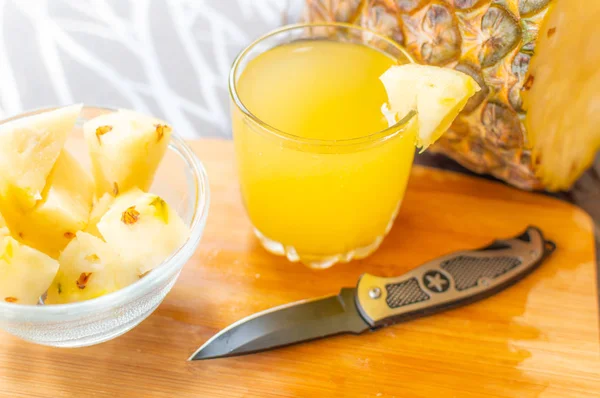 Side view of Sliced pineapple kept on a wooden table besides a glass filled with pineapple juice and a glass bowl with pineapple slices and a knife kept on the chopping board