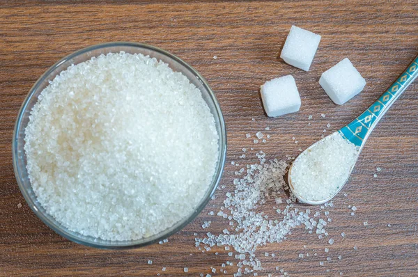 Top view of white sugar kept in a glass bowl with a spoon. Some sugar cubes kept on a clean wooden floor. Sugar taken out from a bowl with a spoon.
