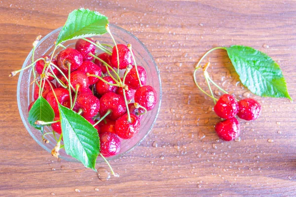 Top view of freshly picked red ripe cherries in a glass bowl kept on a wooden table in kitchen. Some cherries on the wooden table.
