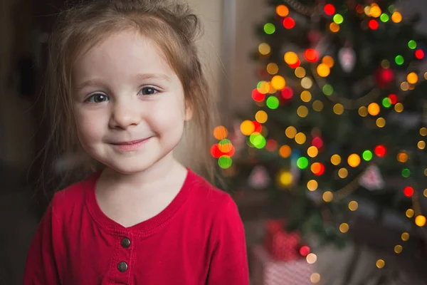 Kırmızı pijamalı tatlı kız Noel ağacının arkasında gülümsüyor. çocuklar ve yeni yıl