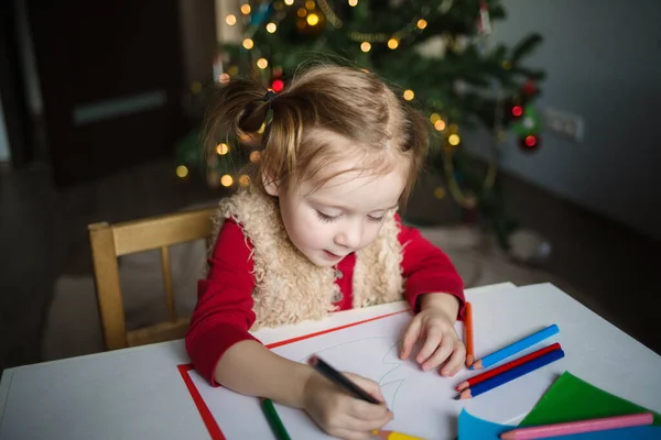 Çocuk Noel Baba 'ya bir mektup yazıyor. Küçük bir kız Noel ağacının arka planına pastel boyalarla resim çiziyor. Çocuklar için yeni yıl yaratıcılığı kavramı.