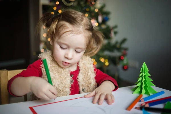 Çocuk Noel Baba 'ya bir mektup yazıyor. Küçük bir kız Noel ağacının arka planına pastel boyalarla resim çiziyor. Çocuklar için yeni yıl yaratıcılığı kavramı.