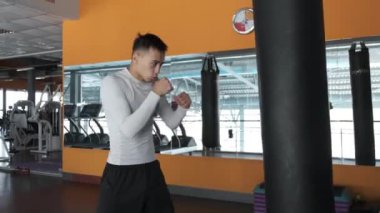 Genç bir Asyalı kickboks spor salonunda eğitim alıyor. Atlet ellerini kum torbasında dövüyor. Aynalı spor salonu.