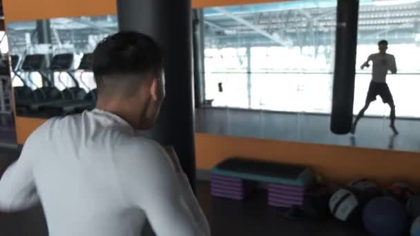 若いアジア人がキックボクシングジムでトレーニングをしています アスリートはパンチバッグで手を叩く ミラー付きジム — ストック動画