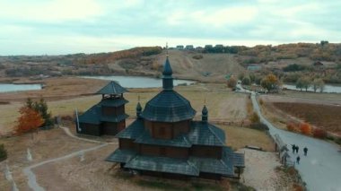 Ahşap kilise ve değirmenlerin hava manzarası. İnsansız hava aracı, eski siyah ahşap bir kilisenin tepesinden havalanıyor ve sonbaharda göle bakan tepelerdeki bir tarlada değirmenler..