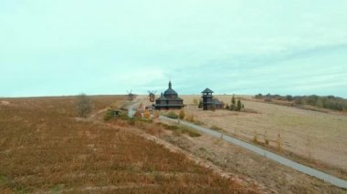 Ahşap kilise ve değirmenlerin hava manzarası. İnsansız hava aracı, eski siyah ahşap bir kilisenin tepesinden havalanıyor ve sonbaharda göle bakan tepelerdeki bir tarlada değirmenler..