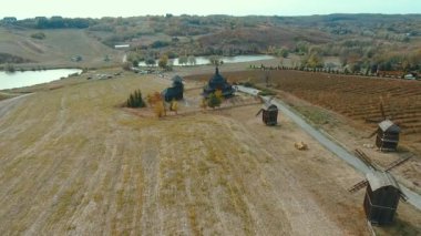 Ahşap kilise ve değirmenlerin hava manzarası. İnsansız hava aracı, eski siyah ahşap bir kilisenin tepesinden havalanıyor ve sonbaharda göle bakan tepelerdeki bir tarlada değirmenler..