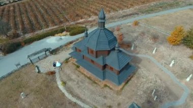 Ahşap kilise ve değirmenlerin hava manzarası. İnsansız hava aracı, eski siyah ahşap bir kilisenin tepesinden havalanıyor ve sonbaharda göle bakan tepelerdeki bir tarlada değirmenler..
