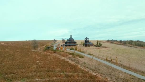 木制教堂和磨坊的空中景观 无人驾驶飞机从一座古老的黑色木制教堂和磨坊的高处起飞 降落在山上俯瞰湖面的田野里 — 图库视频影像
