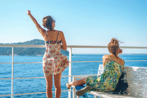 Two young women girl friends sisters sitting on bench fence on deck of the ferry boat or ship sailing to the island tourist destination on summer vacation waving to the horizon in sunny day