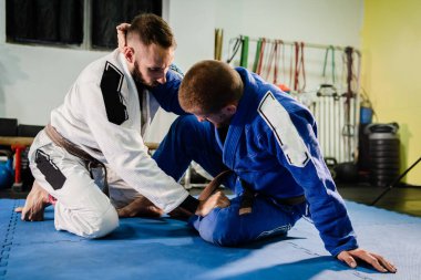 Brezilyalı Jiu Jitsu BJJ dövüş sanatları eğitimi. Akademide iki dövüşçü kelebek pozisyonu almış. Gi kimonosuyla talim teknikleri öğreniyorlar.