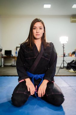 Genç bjj Brezilyalı jiu jitsu kız bayan atlet jujitsu dövüşçüsü tatami paspasların üzerinde eğitim alanında siyah kimono gi ve mavi kemer giyerek oturuyor.