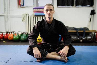 Genç Bjj Brezilyalı jiu jitsu atleti jijitsu dövüşçüsü tatami paspas döşemesinde oturuyor. Eğitimde siyah kimono giysisi ve kemer takıyor.
