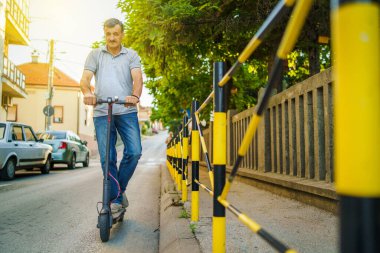 Son sınıf öğrencisi, bir yaz günü, siyah ve turuncu çitli kaldırım kenarında, elektrikli tekme ve scooter kullanıyor.