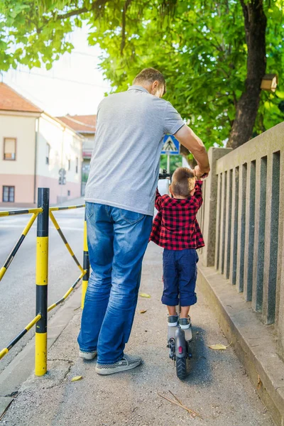 夏の日に町の通りの歩道に乗ってキックスクーターに乗って父や祖父の男と小さな男の子の子供のバックビュー — ストック写真