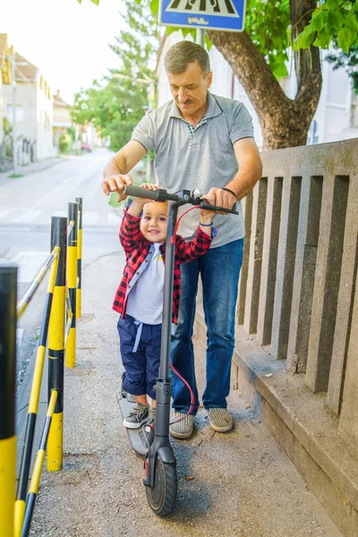 小さな男の子子供オンザ電動プッシュキックスクーターとともに彼の祖父シニア男ヘルプ彼に乗ることを学ぶために歩道 — ストック写真