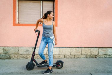 Şehirdeki bir evin duvarının önündeki kaldırımda elektrikli scooter panosunun yanında duran genç bir kadın arkasına bakıyor.