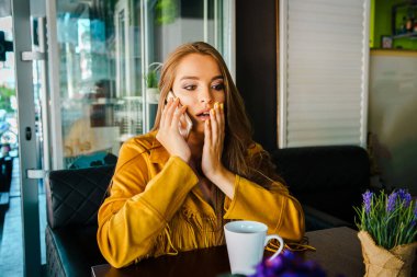Kafe restoranında cep telefonuyla konuşan genç ve güzel bir kadının portresi rahatsız edici bir şekilde şaşkınlık verici bir şekilde el ele tutuşarak tepki veriyor.