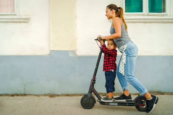 小さな息子や弟の少年を運転して町のいくつかの家の壁の前の舗装上の電動キックスクーターボードに立って若い女性 — ストック写真