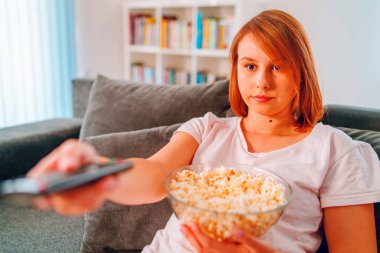 Evdeki kanepede oturan genç bir kadın, elinde patlamış mısır kasesiyle televizyon izleyip evinde tek başına film izleyerek ya da uzaktan kumandayla uzaktan kumandayı tutarak eğleniyor.