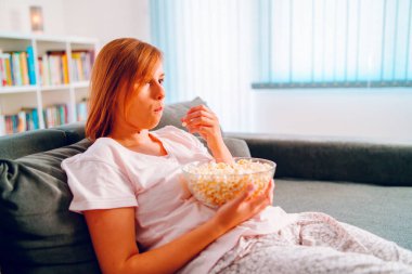 Evdeki kanepede oturan genç bir kadın, elinde patlamış mısır kasesiyle televizyon izleyip evinde tek başına film veya diziden zevk alıyor.