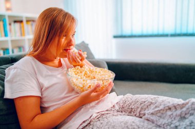 Evdeki kanepede oturan genç bir kadın, elinde patlamış mısır kasesiyle televizyon izleyip evinde tek başına film veya diziden zevk alıyor.