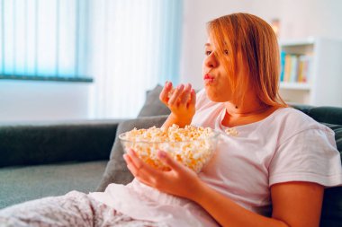 Evdeki kanepede oturan genç bir kadın, elinde patlamış mısır kasesiyle televizyon izleyip evinde tek başına film veya diziden zevk alıyor.