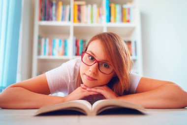 Genç ve güzel bir kız öğrenci, çalışma odasındaki kitap raflarının önünde yerde yatıyor ya da elleri üzerinde kameraya bakarken kameraya bakıyor.