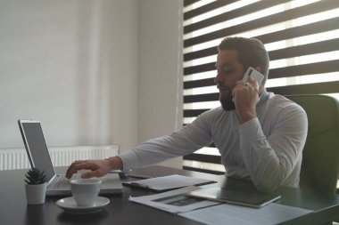 Ofisteki genç adam telefon görüşmesi yapıyor - beyaz iş adamının yan görüntüsü - cep telefonu ve dizüstü bilgisayar kullanarak çalışan beyaz adam yöneticisi - gerçek insan stratejisi ve kararları