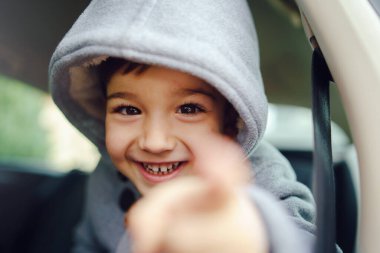 Başında kamerayı işaret eden kapüşonlu çocuk koltuğunda oturan, kışlık ceketli küçük beyaz çocuk portresi.