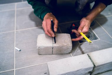 İnşaat işçisi, apartmanın banyosundaki beton tuğla malzemelerini ölçüyor. Ölçüm ölçüsü olarak kalem kullanıyor.