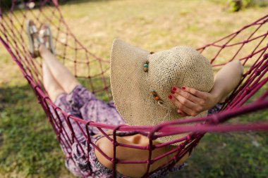Genç bir kadının yaz günlerinde giydiği elbise ve şapkayla arka bahçesindeki hamakta kestirirken görüntüsü.