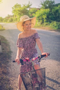 Bir yaz günü, güneşli bir günde açık yol asfaltında elbise ve şapka giyip bisiklete binen genç bir kadın.
