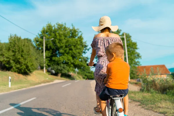 若いです女性で晴れた夏の日に身に着けていますドレスに乗って自転車に乗ってアスファルトの上の道路上で彼女の小さな男の子子供の息子で後ろに座って — ストック写真