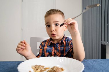 Küçük beyaz bir çocuğun portresi masaya oturmuş öğle veya akşam yemeği yiyor çatal tutuyor ve yüz ifadeleri yapıyor.