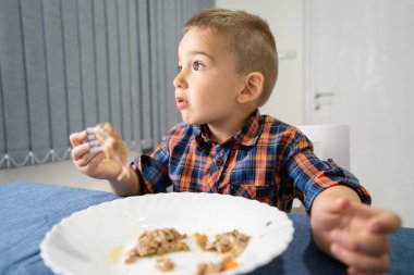 Küçük beyaz çocuk portresi. Masada oturmuş öğle veya akşam yemeği yiyor. Çatal tutuyor.
