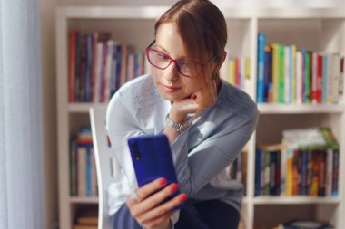 Kafkasyalı genç bir kadın kadın, evinde sandalyede oturuyor ya da kitaplığın önünde elinde akıllı bir cep telefonuyla çalışıyor. Selfie çekiyor ya da gündüz görüşünde video çekiyor.
