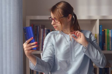 Genç, beyaz, kadın, evinde sandalyede oturuyor ya da kitaplığın önünde elinde cep telefonuyla çalışıyor. Selfie çekiyor ya da gündüz görüşünde bir video görüşmesi yapıyor.