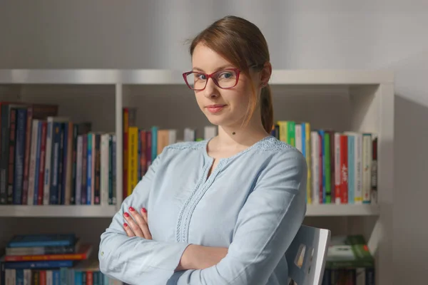 Waist up portrait of young caucasian woman adult girl sitting on the chair at home or office in front of the book shelf wearing glasses and shirt looking to the camera front view in morning day