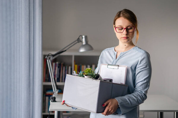 Young adult caucasian woman female girl standing by the window at the office losing her job holding personal items things in box being fired from work dismissed due to crisis recession quitting