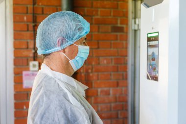 İşe gitmeden önce dijital sıcaklık kontrol kamerasıyla elektronik yüz tanıma terminalinin önünde duran beyaz doktor kıyafetleri giyen bilim adamının yan görüntüsü.