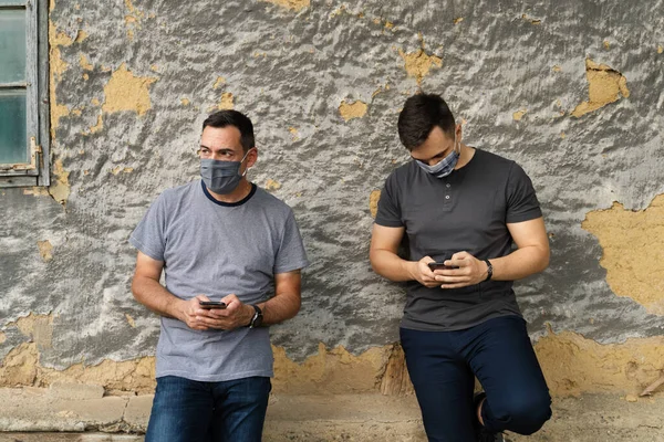 两名男子在关门大吉后戴着防护面具在墙前使用手机应用程序 旅行或度假时通过互联网保持社交距离 新的正常生活方式概念 — 图库照片