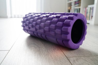 Bulanık mobilyaların önündeki açık beyaz ahşap zeminde vücut masajı için mor desenli köpük silindiri.