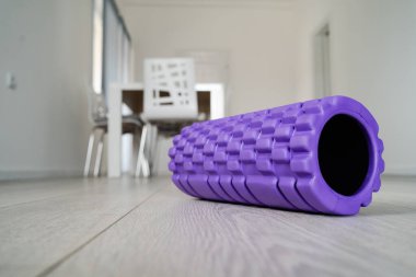 Bulanık mobilyaların önündeki açık beyaz ahşap zeminde vücut masajı için mor desenli köpük silindiri.