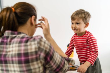 Küçük beyaz bir çocuğun omuzlarının üzerinden annesinin önünde duran ve evde masanın başında oynayan küçük beyaz bir çocuk.